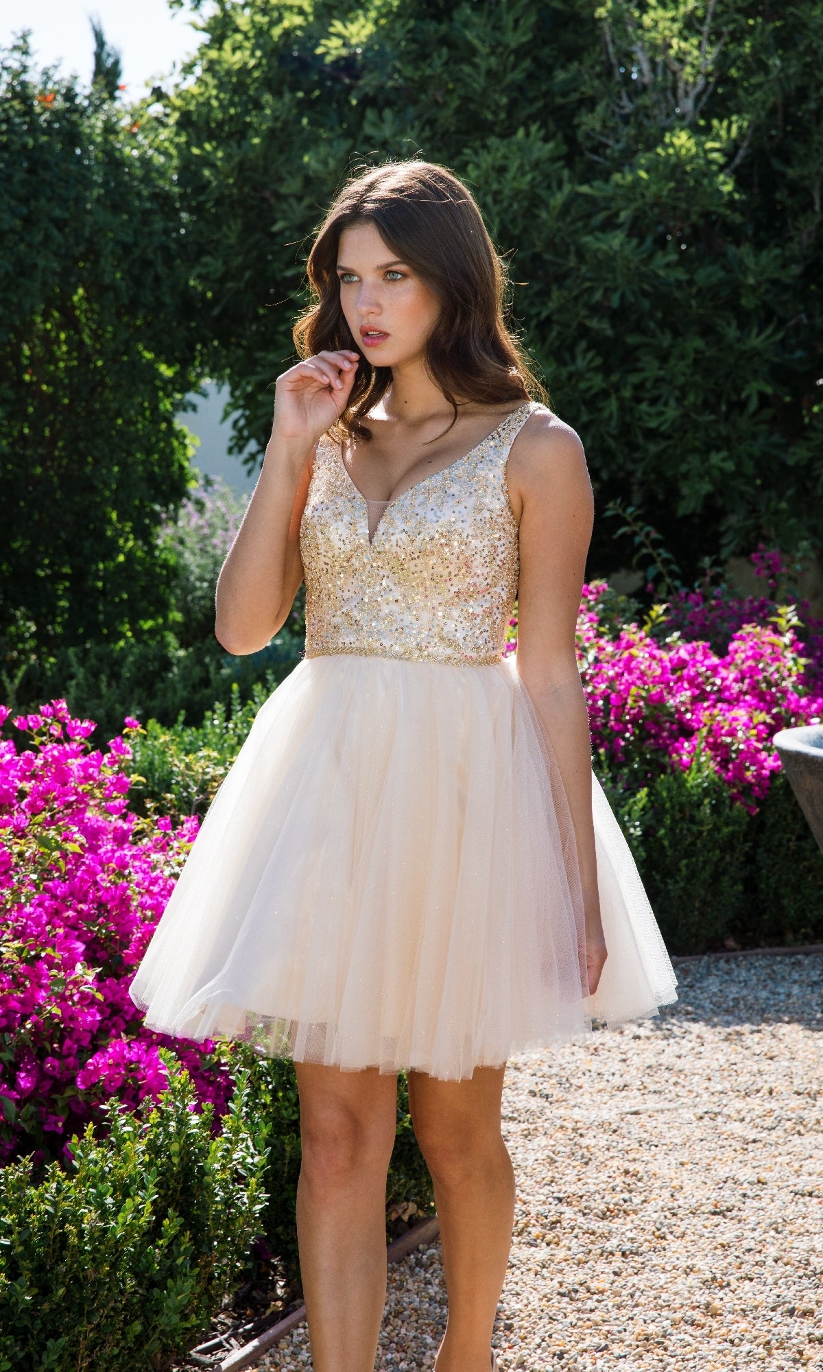 A woman in the Eureka 9926 Glitter-Bodice Short A-Line Hoco Dress with a gold sequin bodice and tulle skirt stands outdoors near blooming pink flowers and lush greenery.